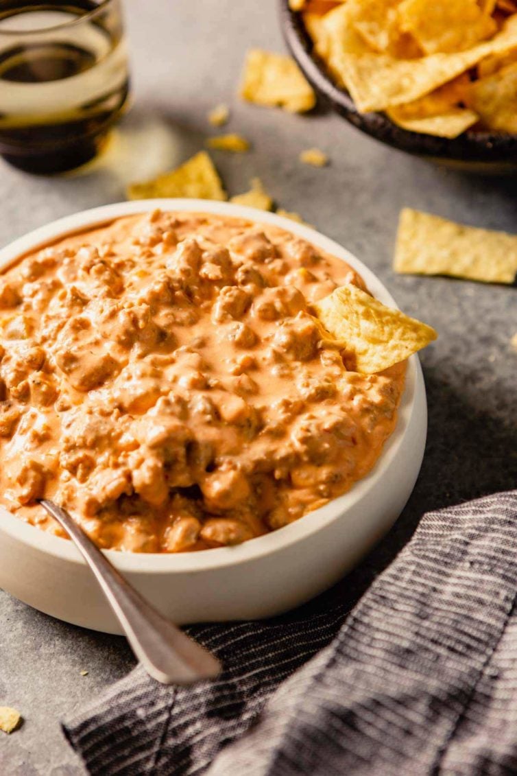 A white bowl of sausage dip on a grey counter with a spoon in the left side of the bowl and tortilla chips on the back of the counter.