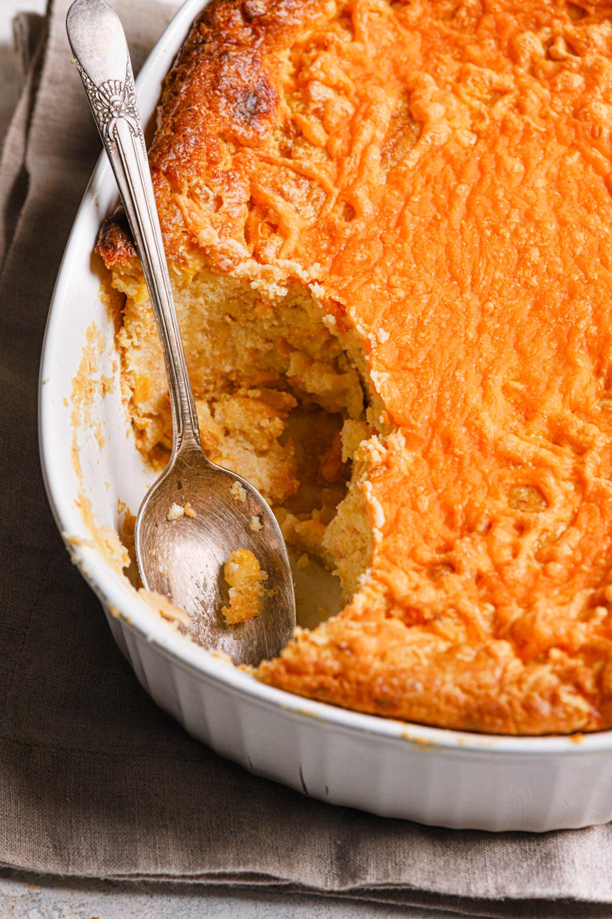 a oval casserole dish filled with thsi baked cheesy creamed corn casserole with a spoon scooping out one serving