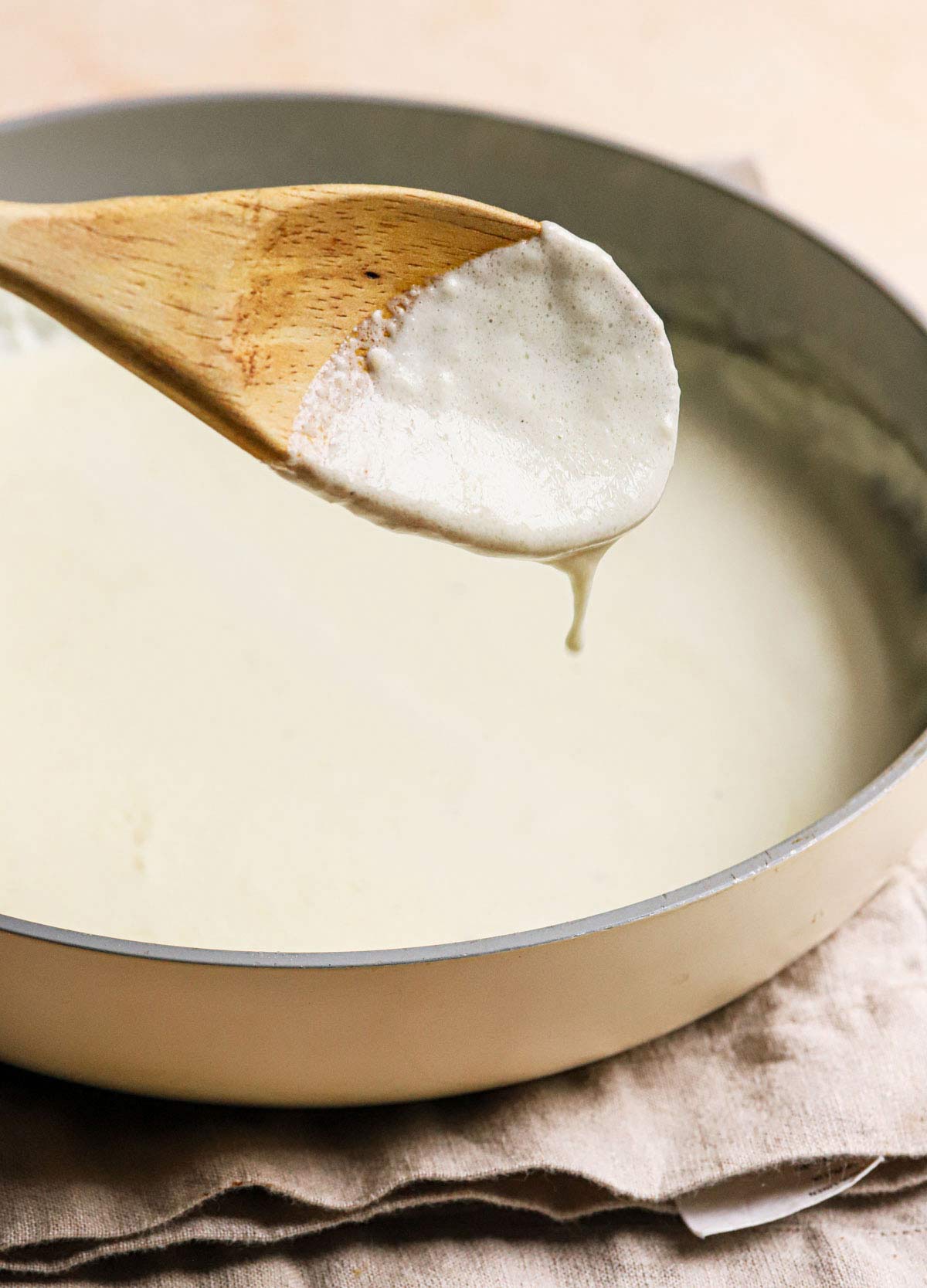 Alfredo sauce dripping off a wooden spoon being held above a saucepan.