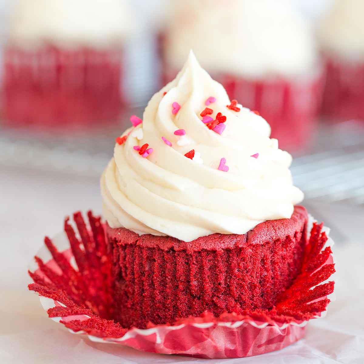 Red velvet cupcake with liner partially removed, cream cheese icing on top with red and pink heart sprinkles.