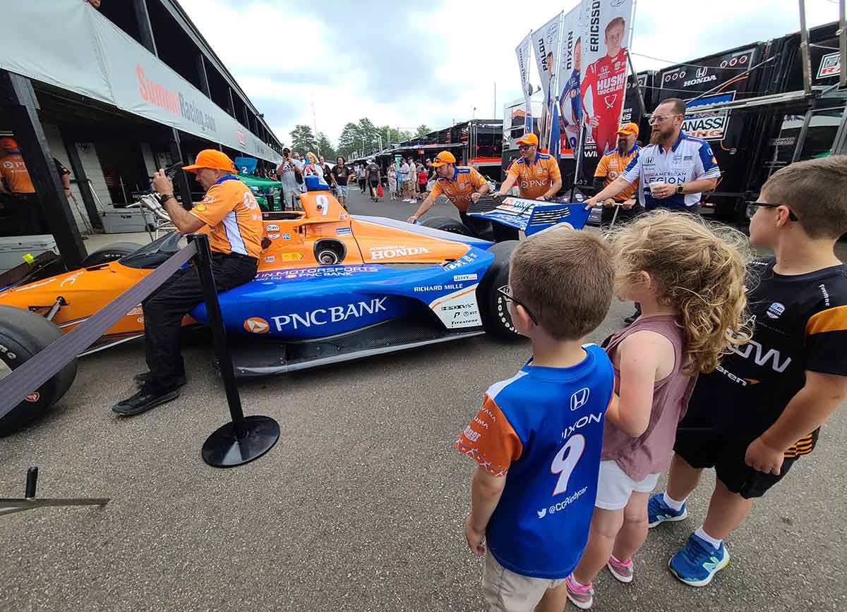 Three kids watching a race car being pushed into a garage area.