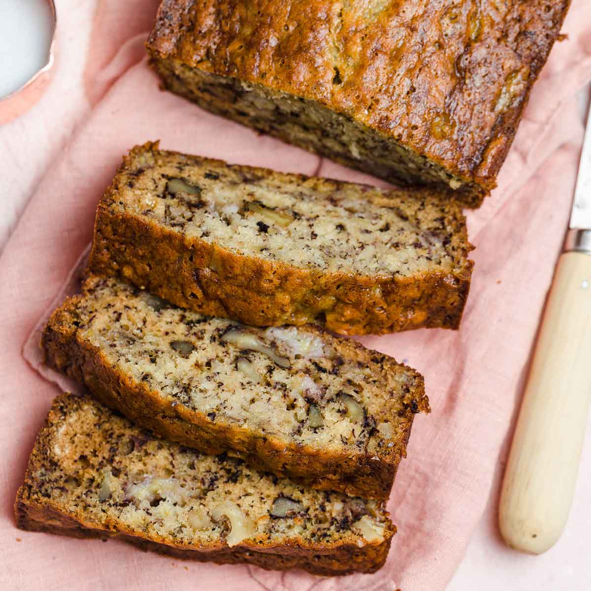 Three slices of banana nut bread laying on a pink cloth napkin in front of the remaining loaf.