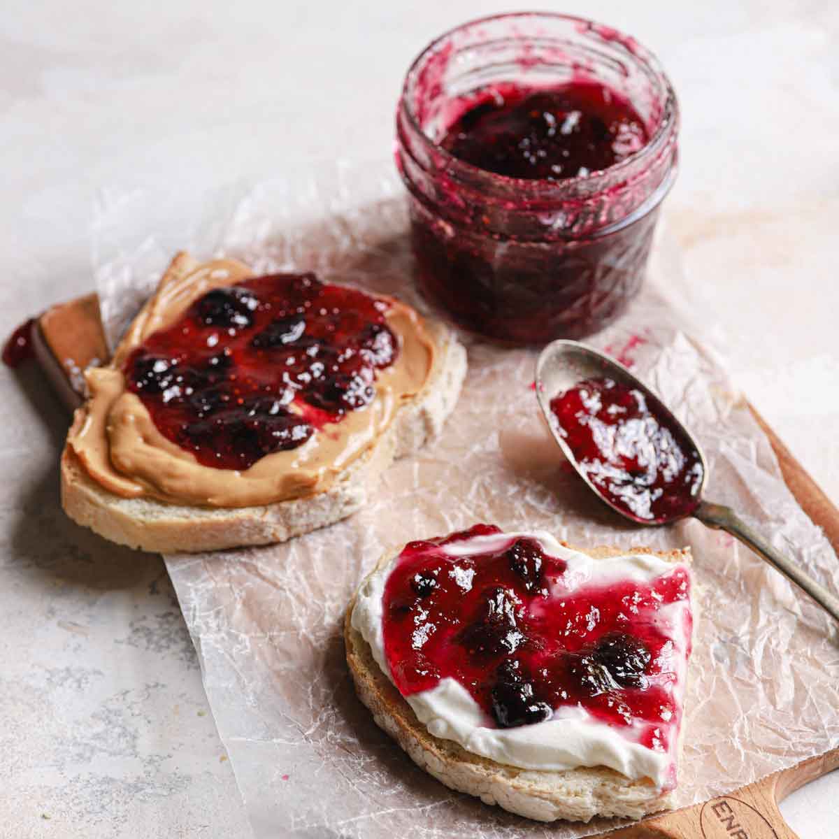 Toast with peanut butter and blueberry jam, a jar and spoon on wax paper on a cutting board.