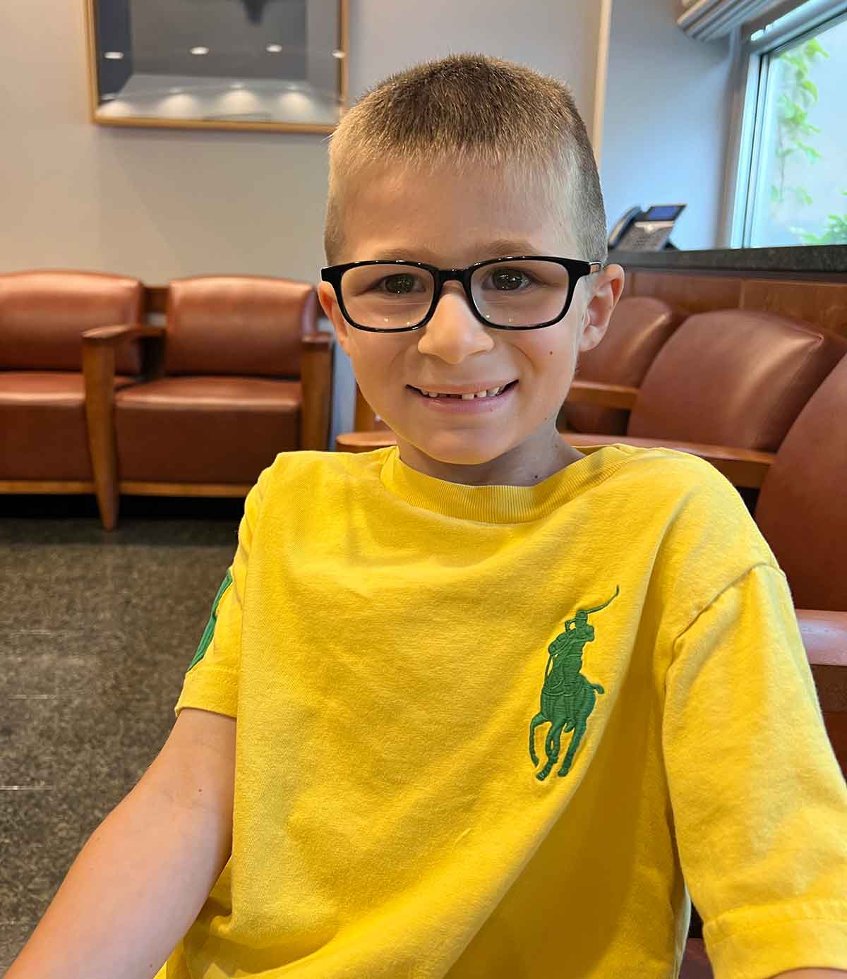 Boy in yellow shirt and black glasses smiling for a photo in a waiting room.