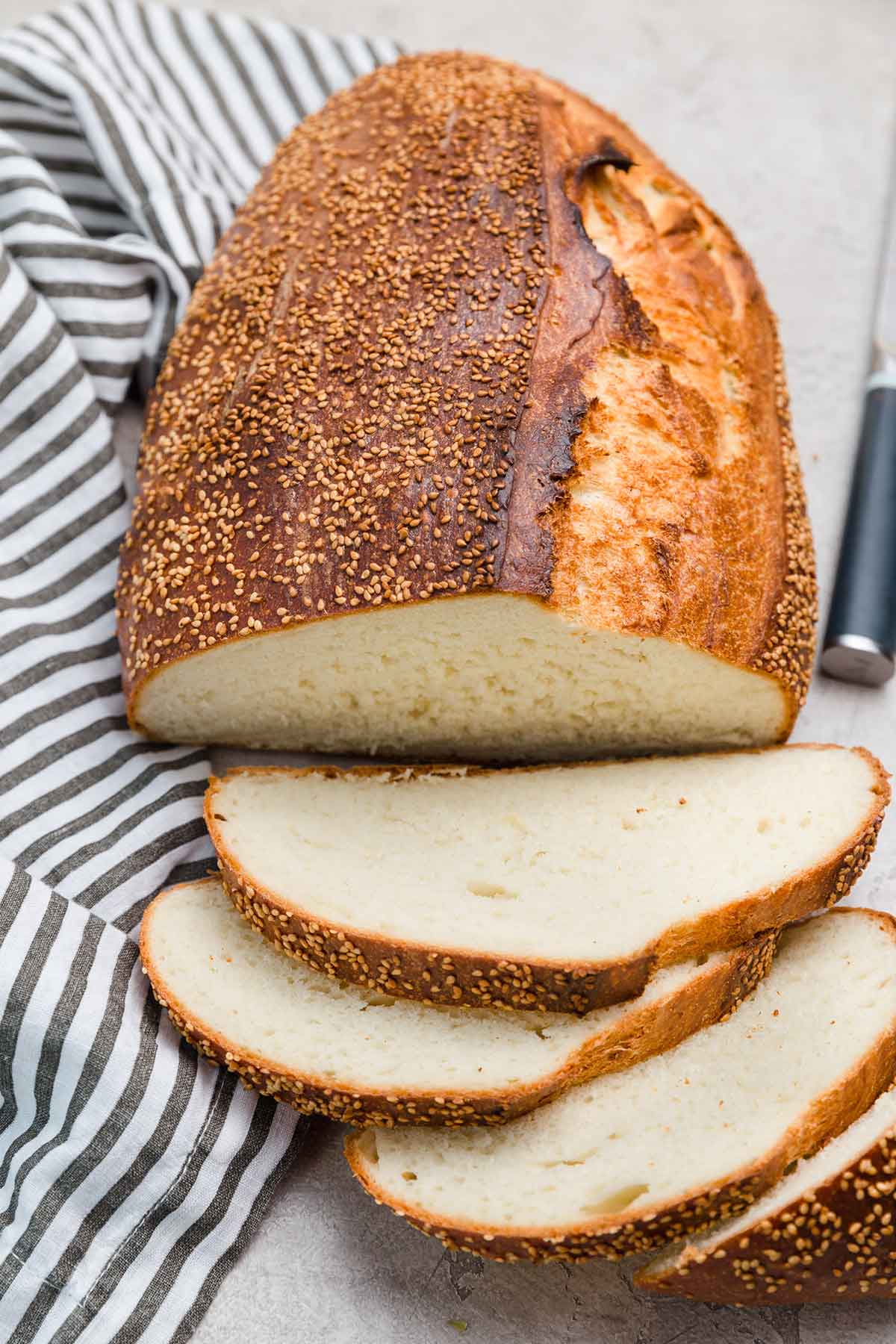Loaf of Italian bread with sesame seeds on top, with four slices sliced off and laying in front of loaf with napkin and knife to the side.