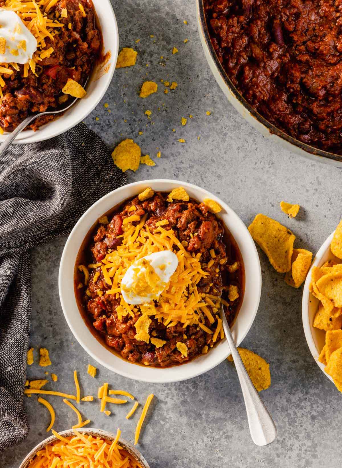 Overhead photo of bowl of chili with cheese, sour cream, and corn chips as toppings on gray countertop.