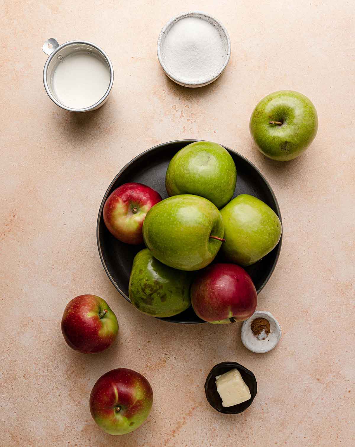 A black plate with a pile of apples on it and on the counter next to it, with bowls of sugar, seasoning, and butter close by.