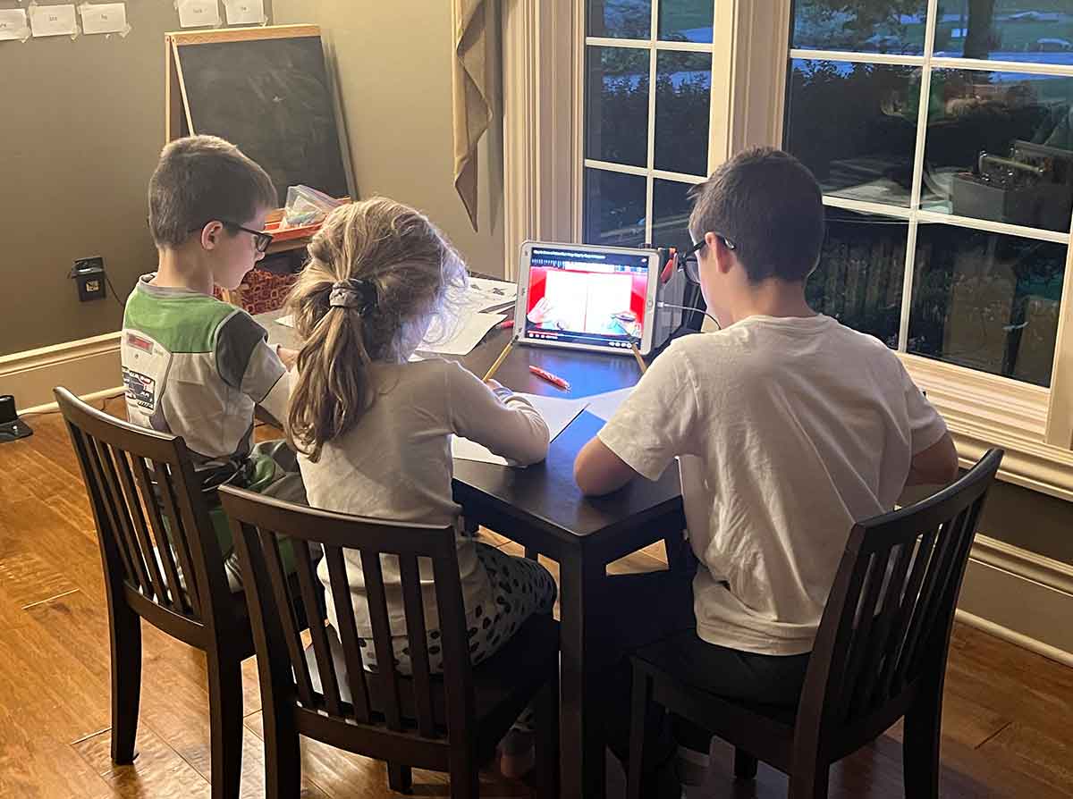 Three kids sitting at a table in front of a window drawing on paper.