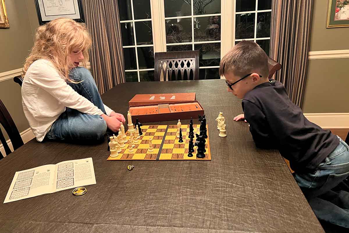 A girl and boy sitting on either side of a chess board at a dining room table.