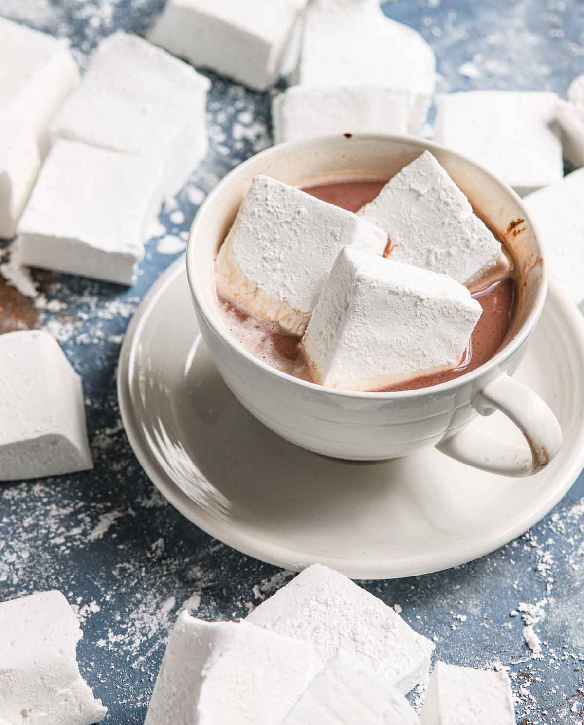 A cup of hot chocolate in a white cup on a white saucer, with three large homemade marshmallows floating on top.