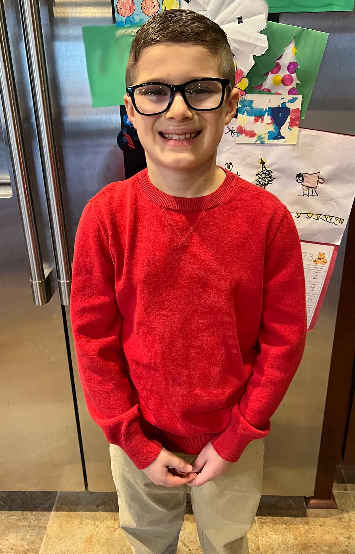 Boy in khaki pants and a red sweater standing in front of a refrigerator.