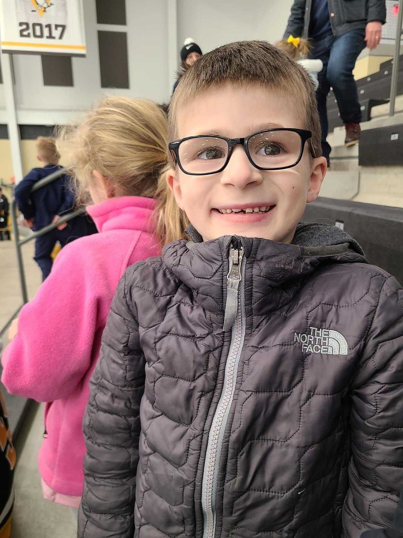 Little boy in a grey winter jacket sitting in bleachers at an ice arena.