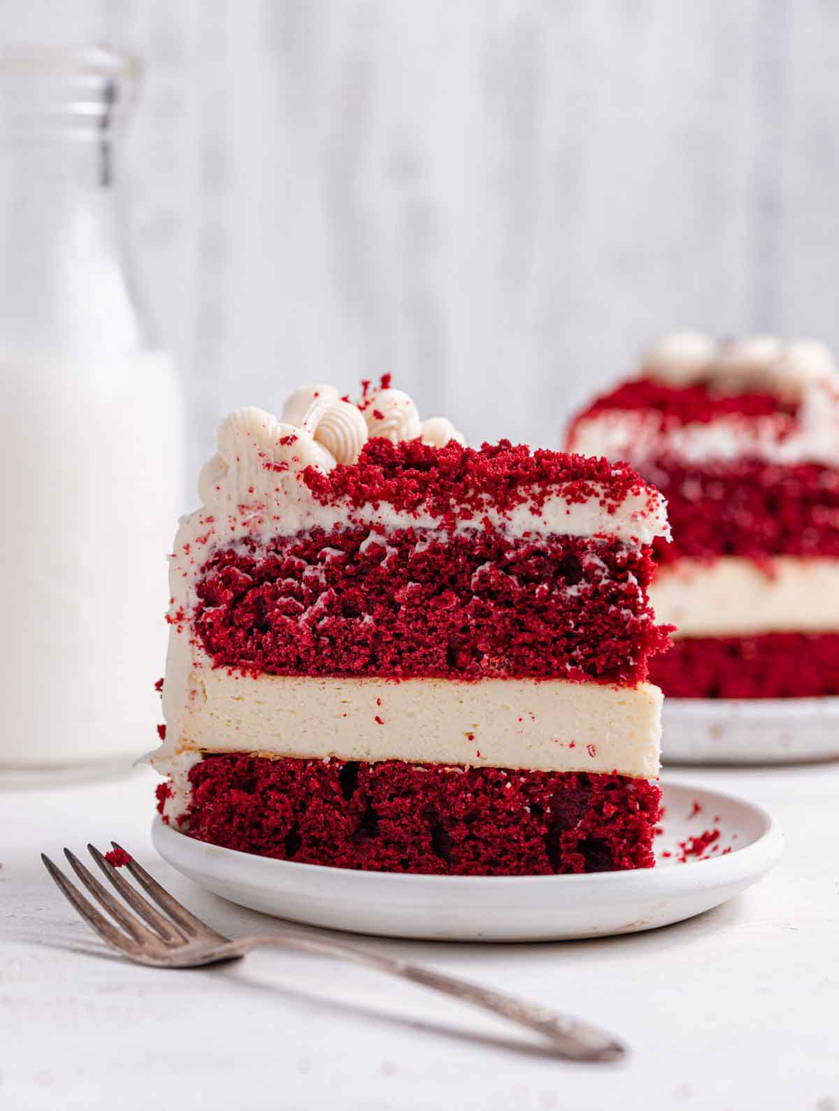 Slice of red velvet cheese on a white plate with a fork in the foreground and another slice in the background.