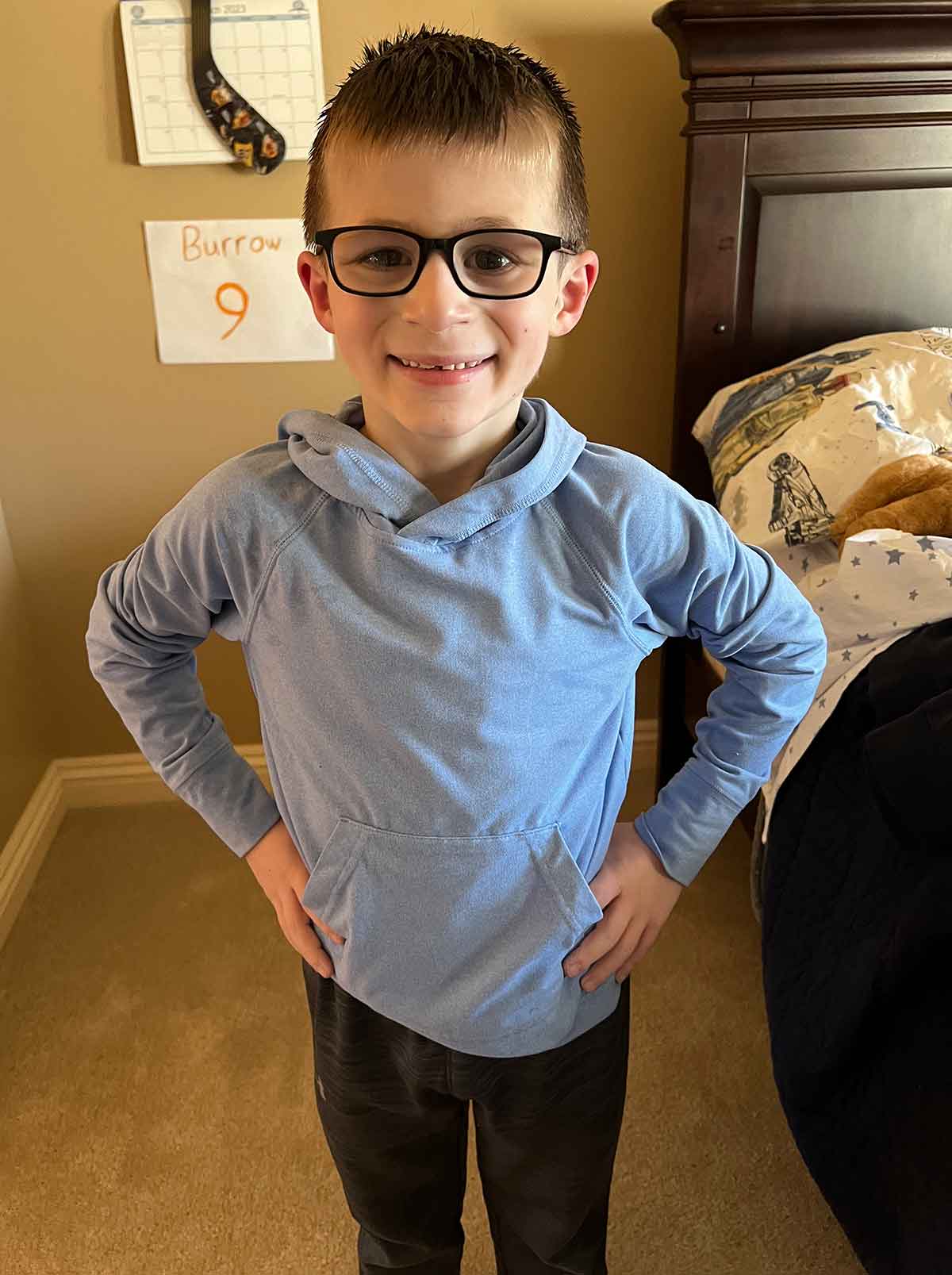 Boy in a blue sweatshirt standing with his hands on his hips in a bedroom.