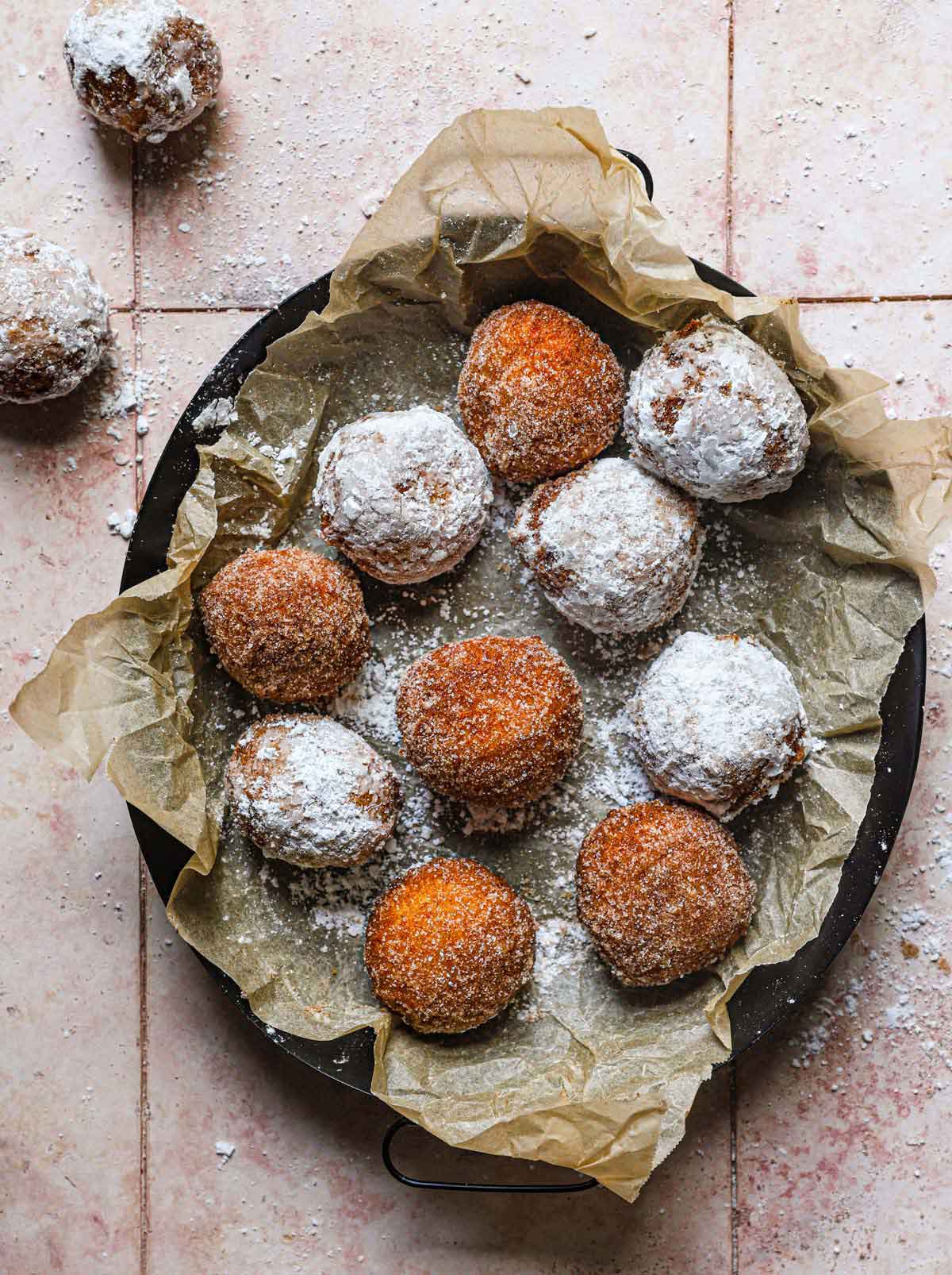 Overhead photo of fried zeppole coated in powdered sugar and cinnamon sugar in parchment paper in a dark pan.