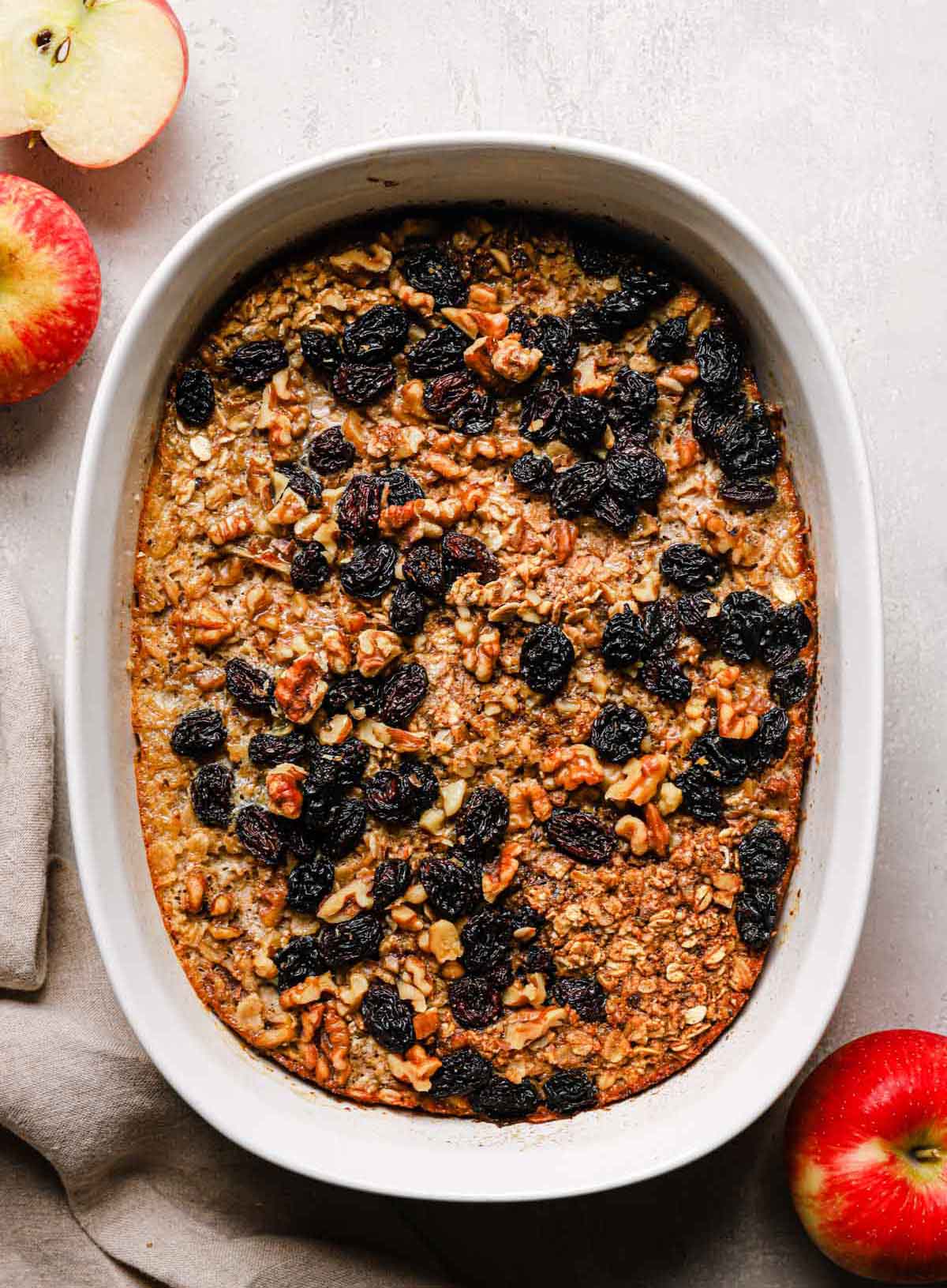 Overhead photo of baked oatmeal topped with raisins and walnuts in white baking dish with cut apple on the side.