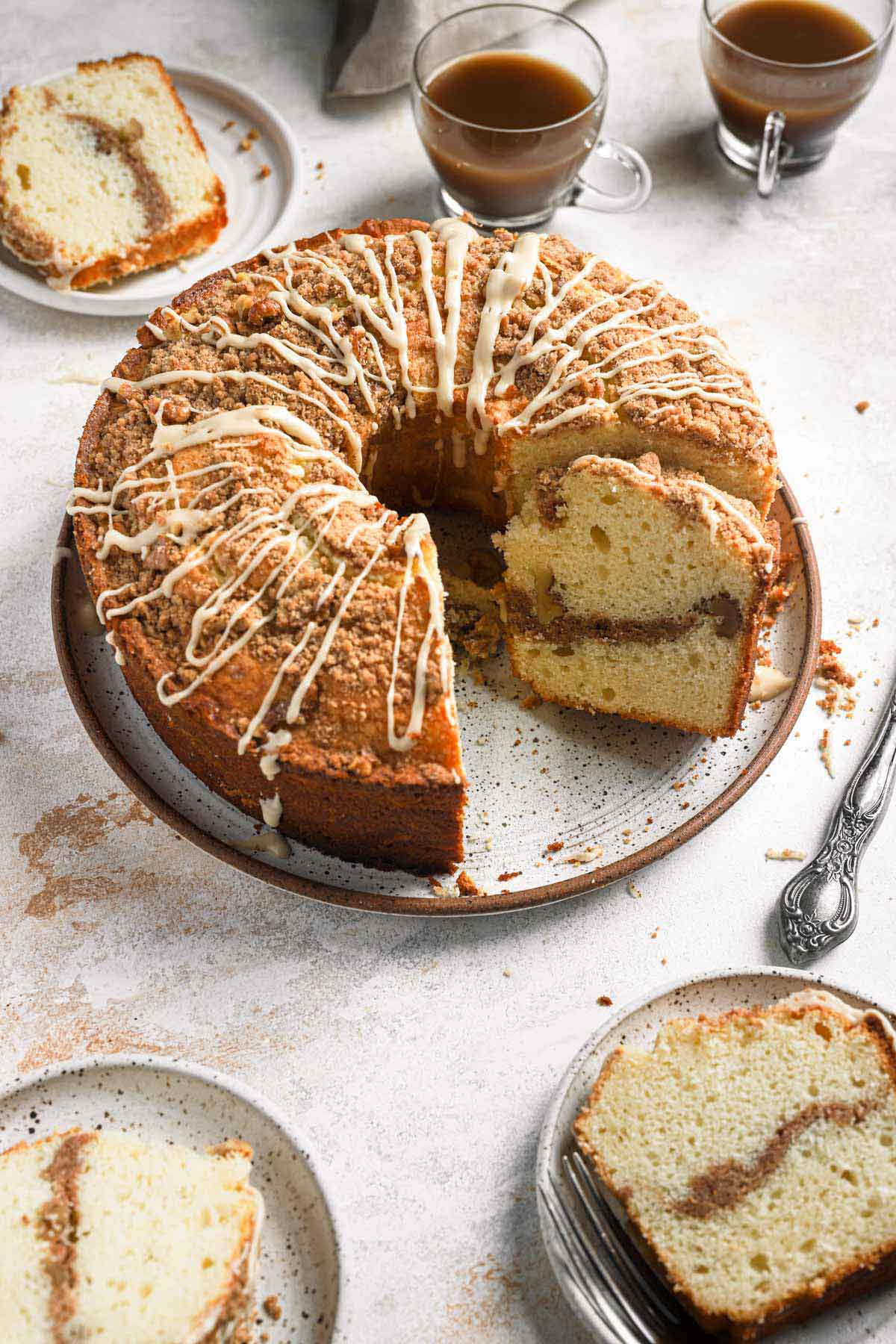 A round coffee cake on a serving plate with one slice cut away and additional slices on plates behind and in front, with two cups of coffee behind the cake as well.