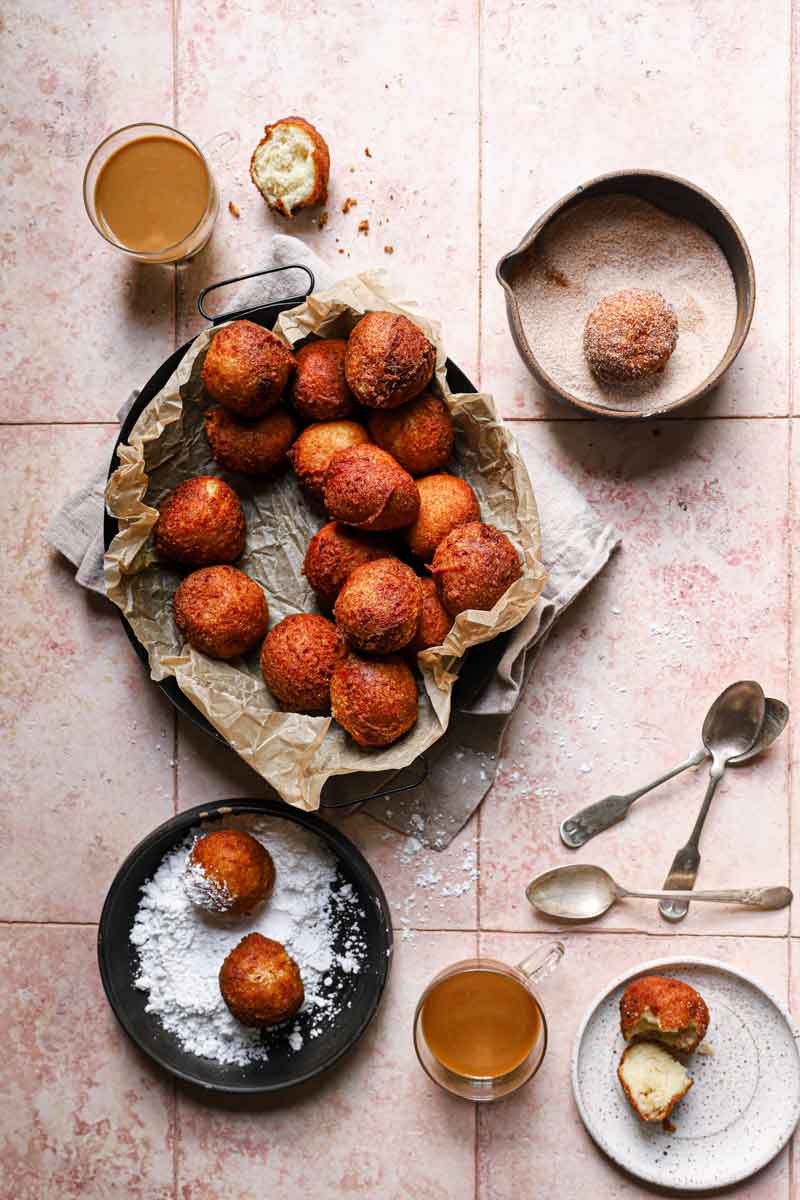 Overhead photo of fried zeppole in parchment-lined pan with some dropped in a bowl of cinnamon sugar and others being rolled in powdered sugar.