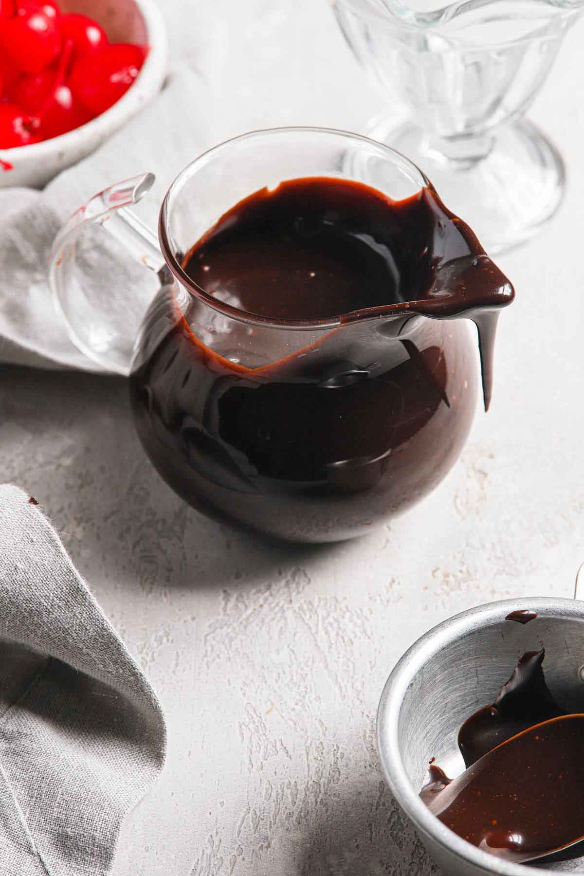 A small glass pitcher filled with hot fudge, with a bit of sauce dripping off the top.