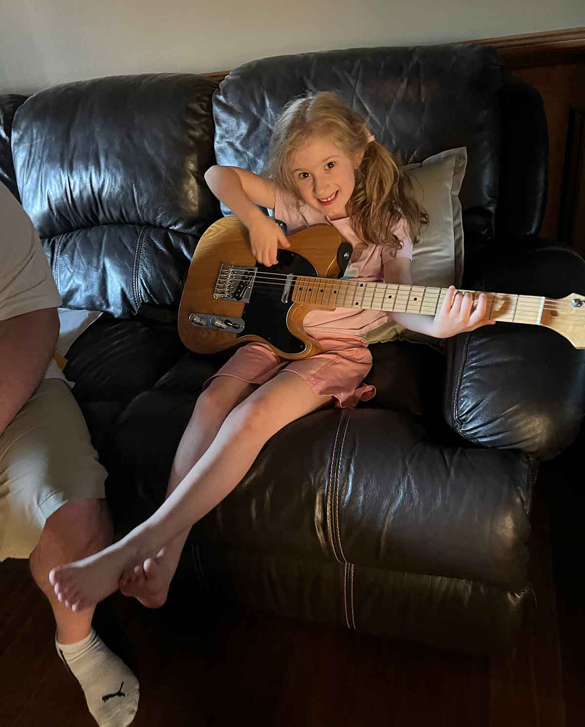 Little girl sitting on a brown leather couch holding a guitar.