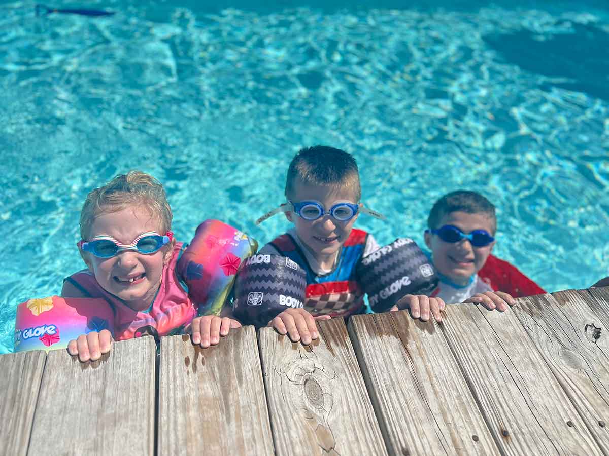 A girl and two boys in a swimming pool, hanging on to the edge, wearing goggles and smiling.