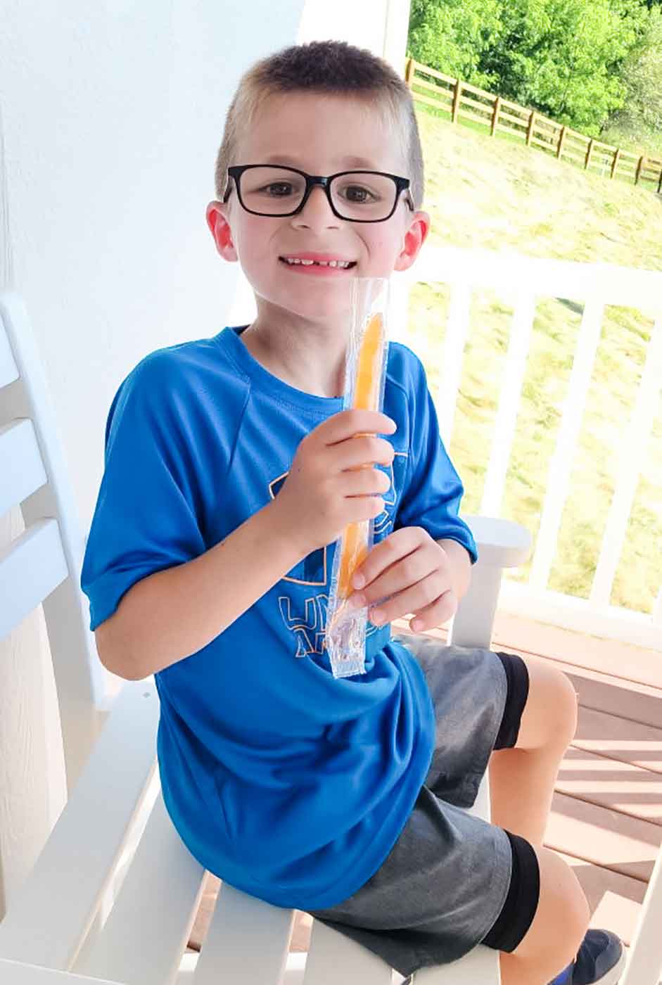 Little boy sitting on a chair eating a popsicle on a deck.