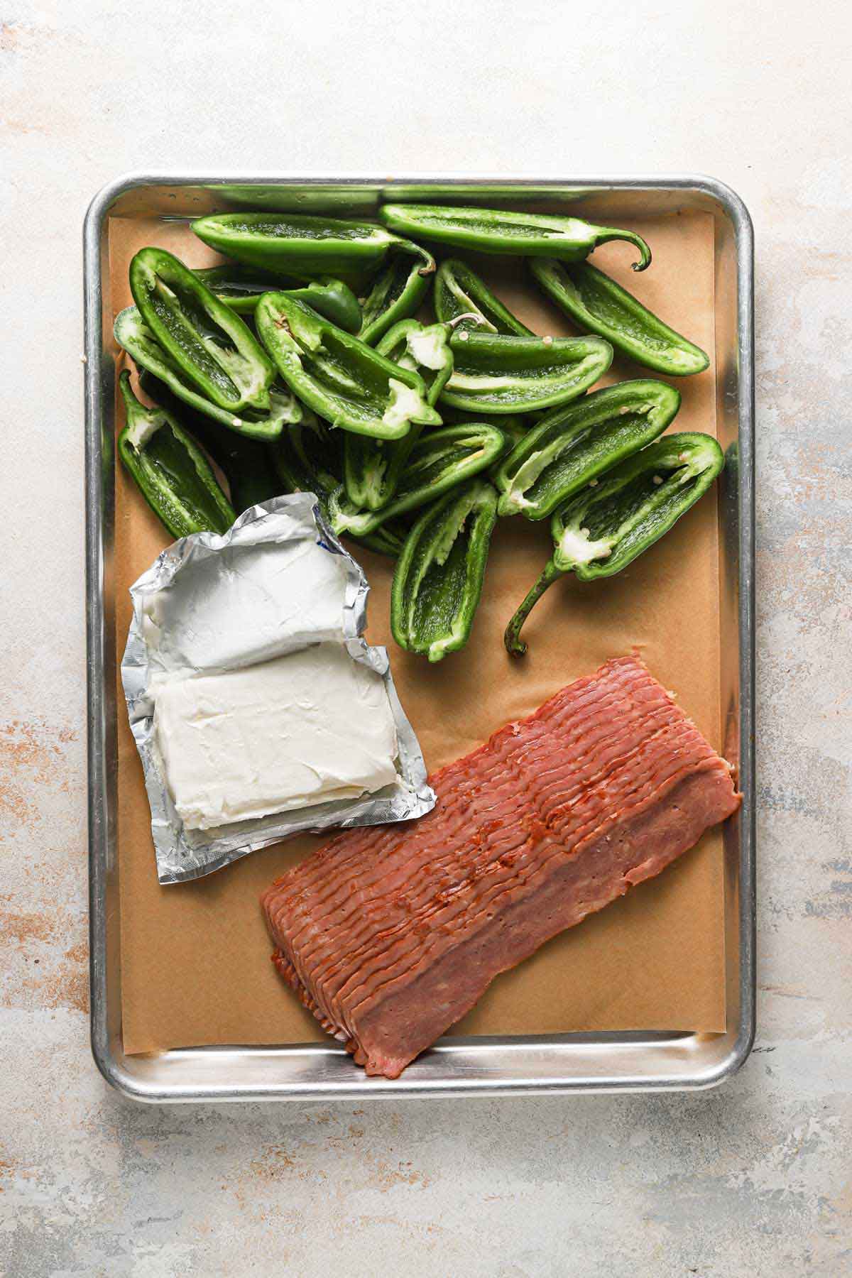 Baking sheet covered in parchment paper with halved jalapeno peppers, cream cheese, and bacon laid out on top.