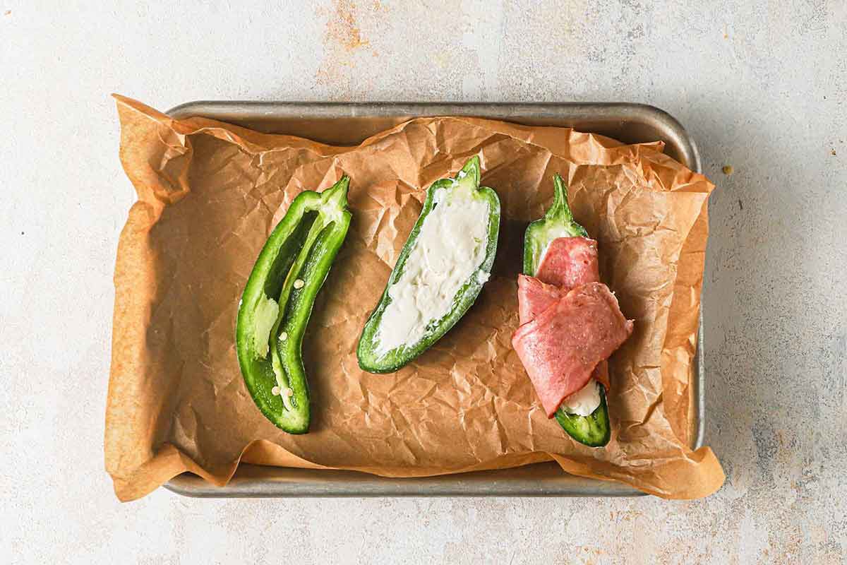 Three jalapeno jalves on parchment paper - one empty, the next one filled with cream cheese, and the last one wrapped in bacon.