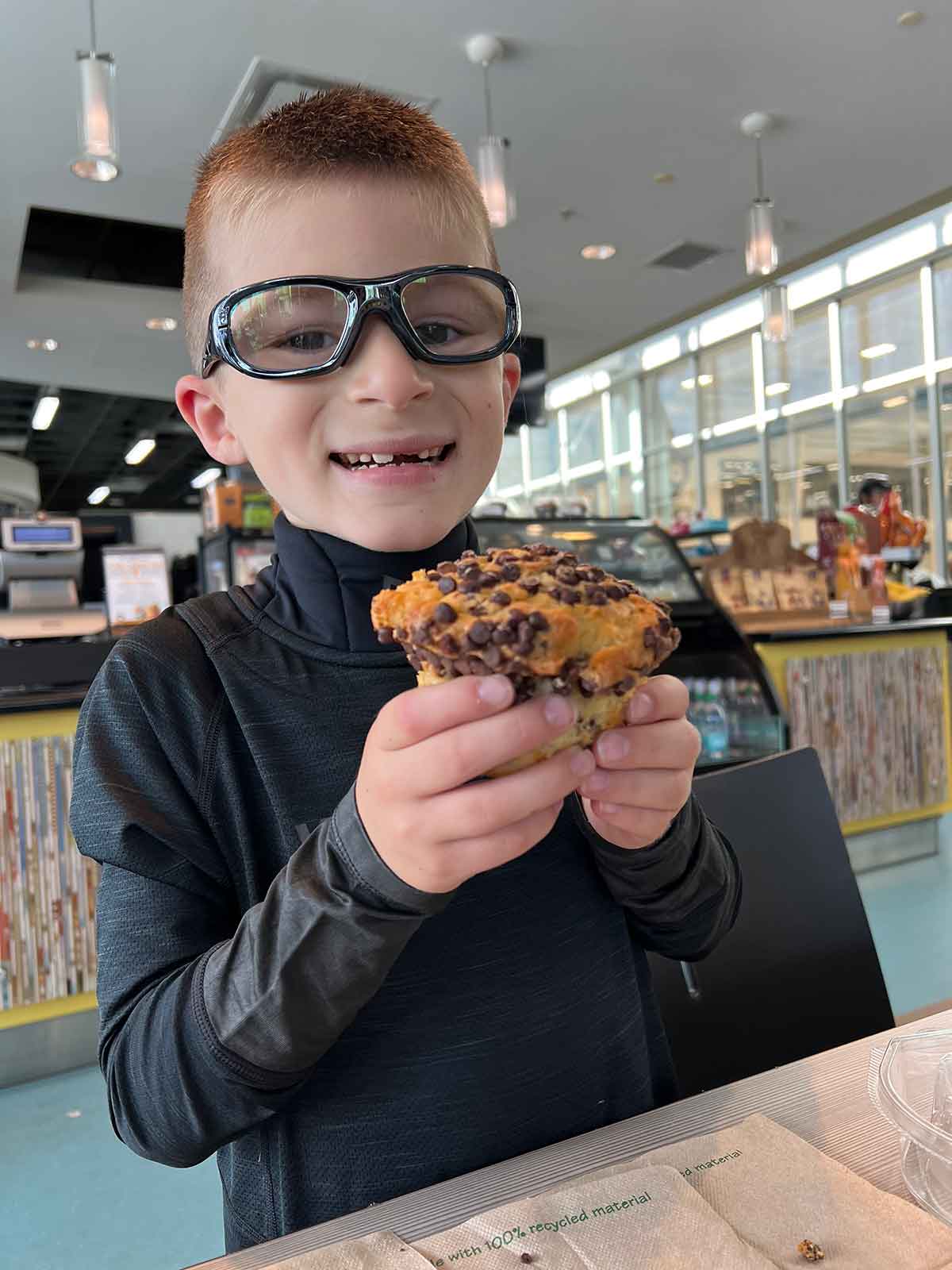 Little boy in black rec specs and hockey base layer eating a chocolate chip muffin.