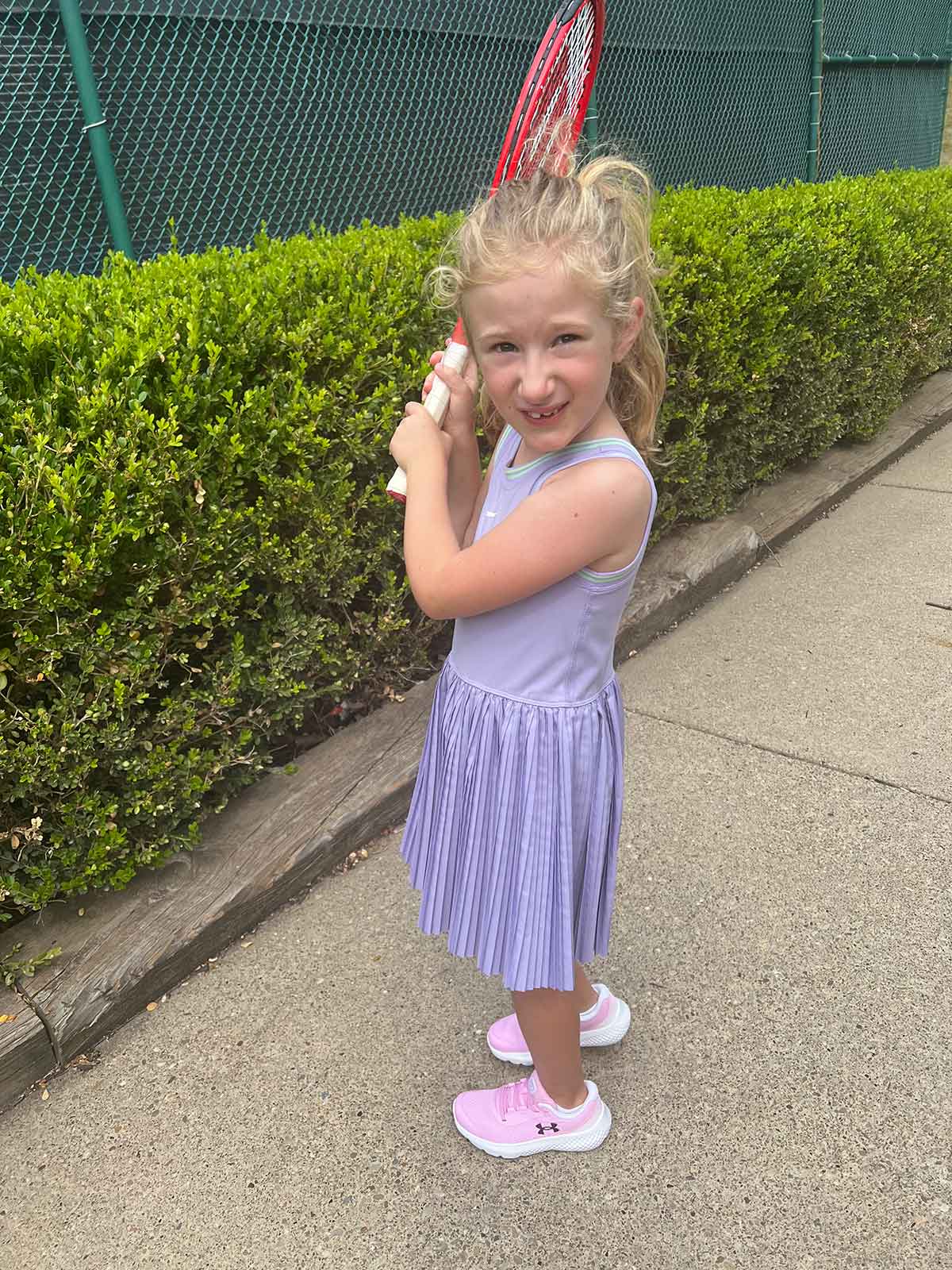 Little girl in purple tennis dress and pink sneakers holding a tennis racket.