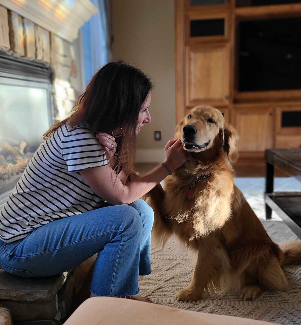 Woman sitting in front of a fireplace petting a Golden Retriever sitting in front of her.