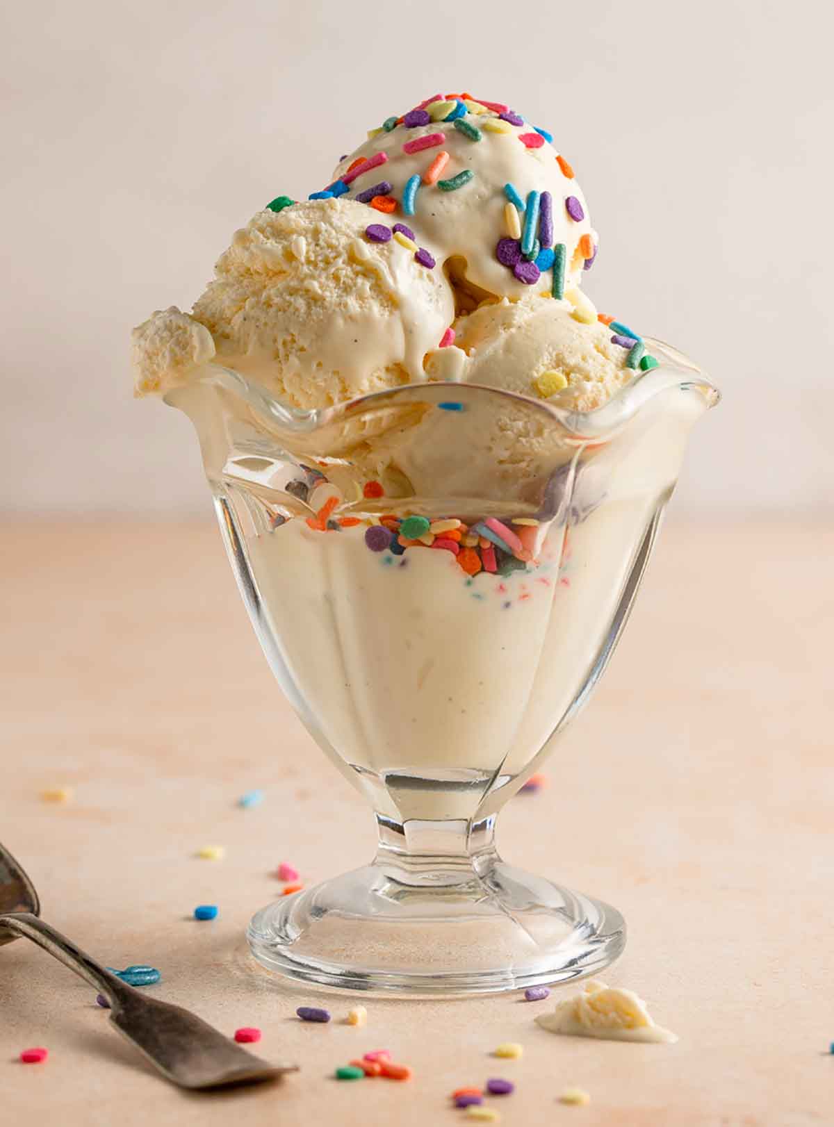Three scoops of vanilla ice cream in a glass parfait dish with colorful sprinkles on top.