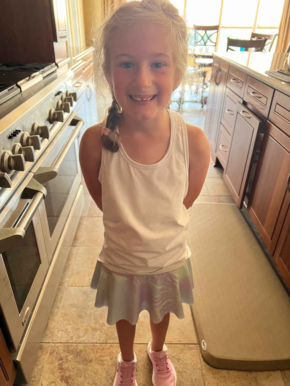 Little girl in white tank top standing smiling in a kitchen.