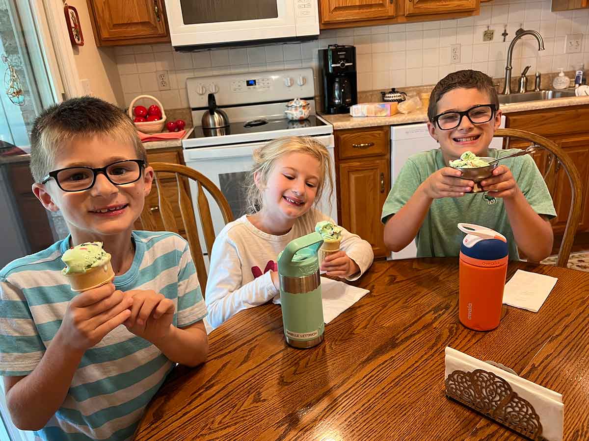 Three kids sitting at a kitchen table eating ice cream cones.