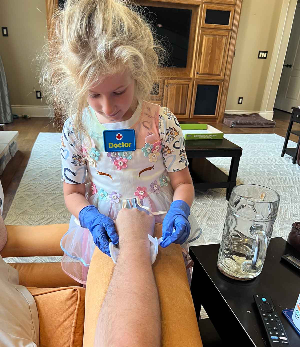 Little girl in pajamas and a unicorn dress playing doctor with gloves and wrapping a pretend bandage around an arm.