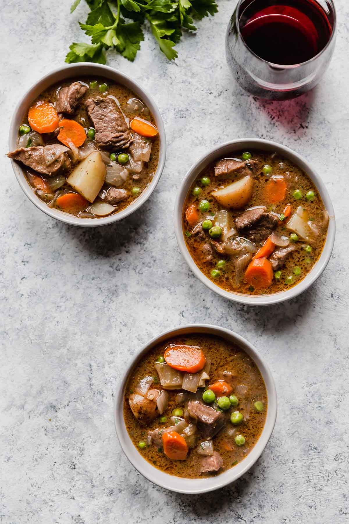 Three small bowls of beef stew arranged on a grey counter, with a glass of red wine in the background.