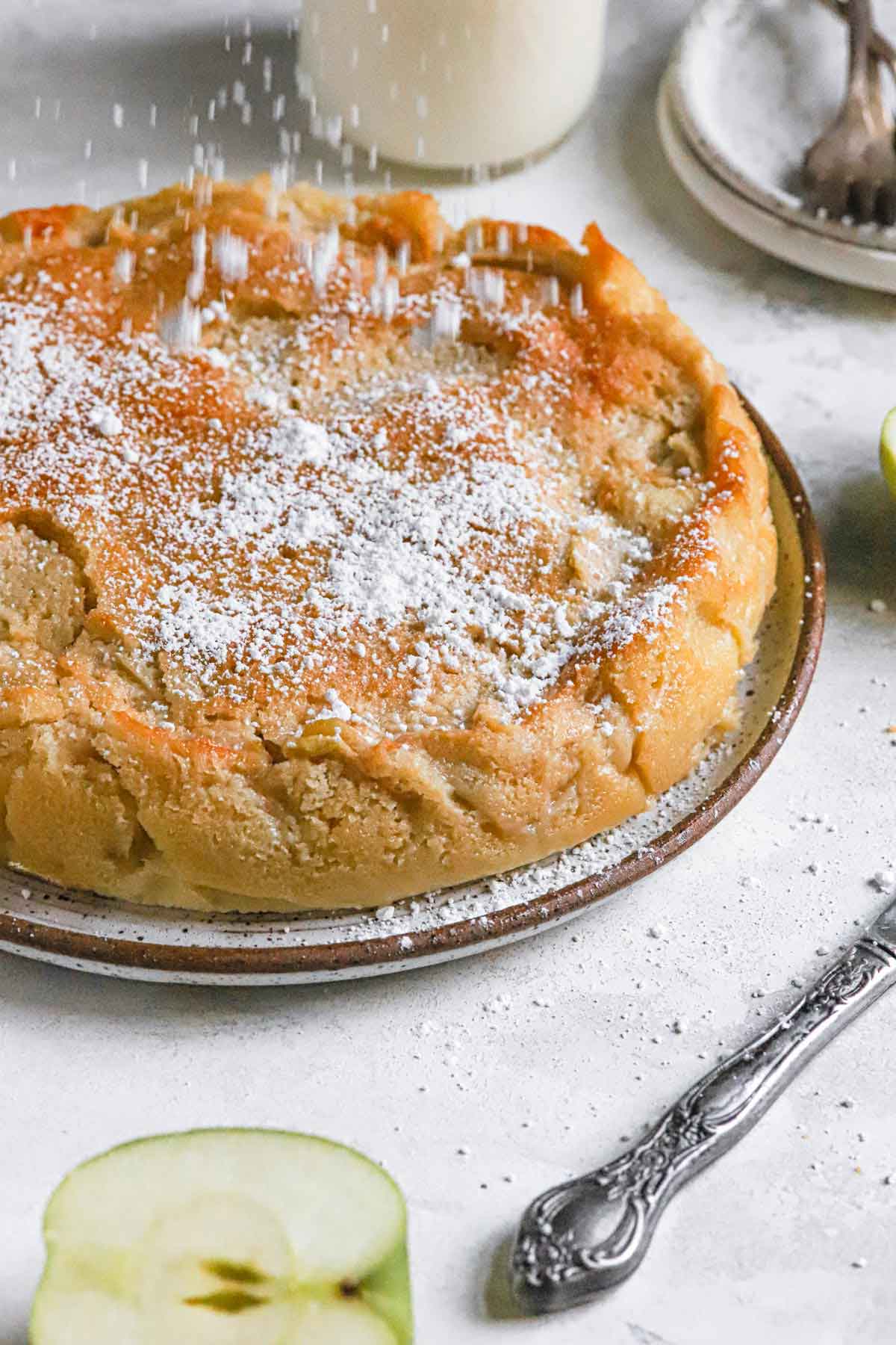 French apple cake on a serving plate being sprinkled with powdered sugar.
