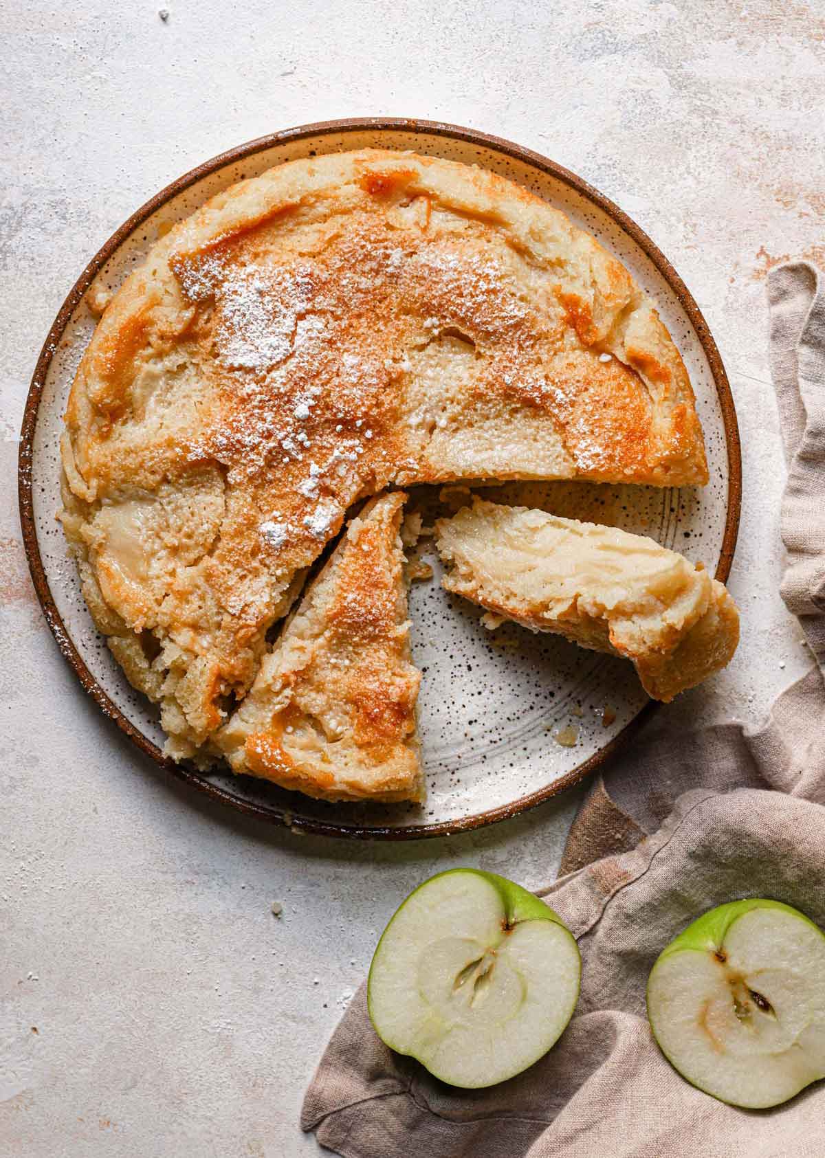 Overhead photo of French apple cake on a serving plate, with one slice cut away and on its side.