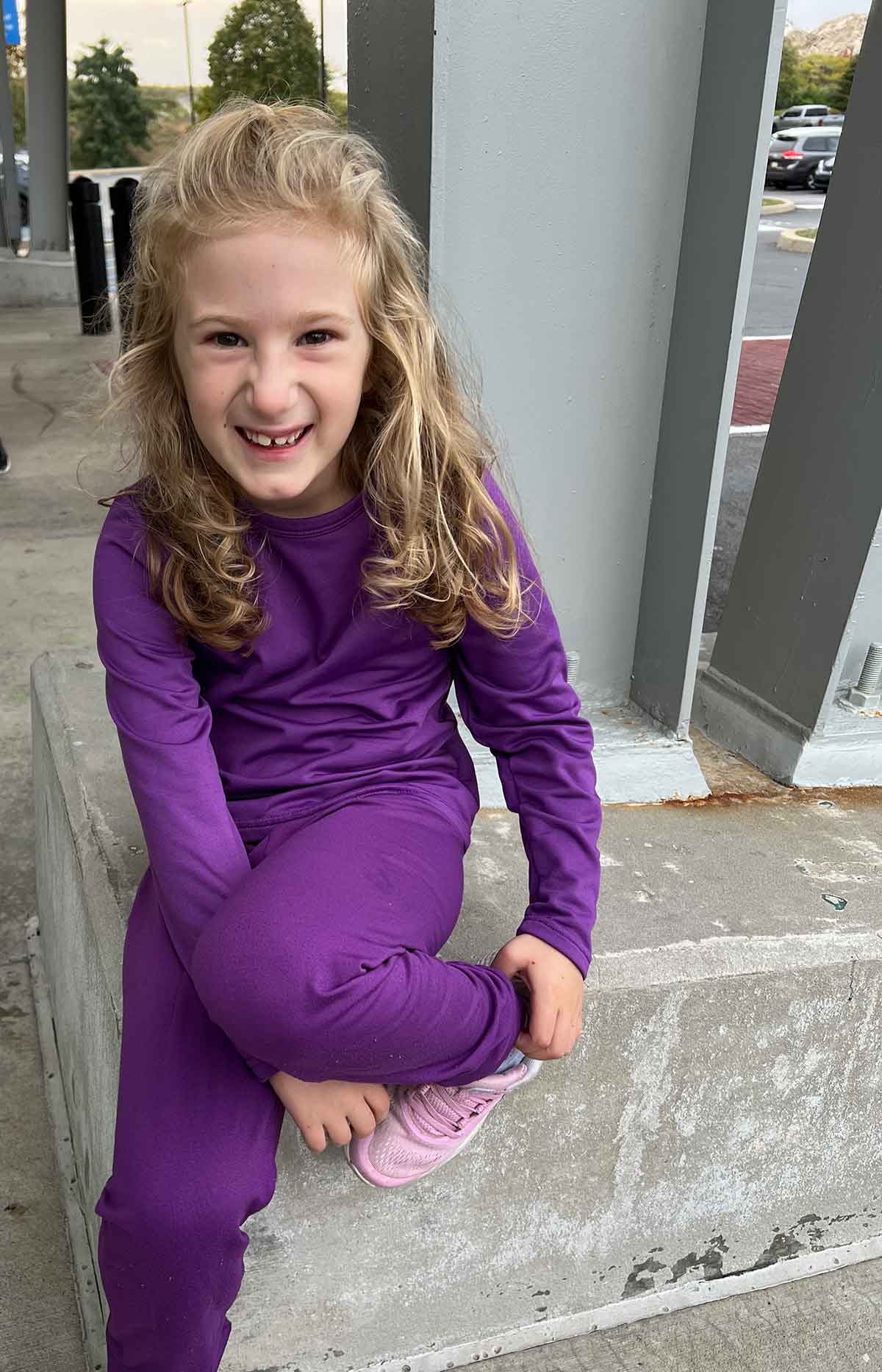 Little girl with blonde hair in a purple sweat suit sitting outside.