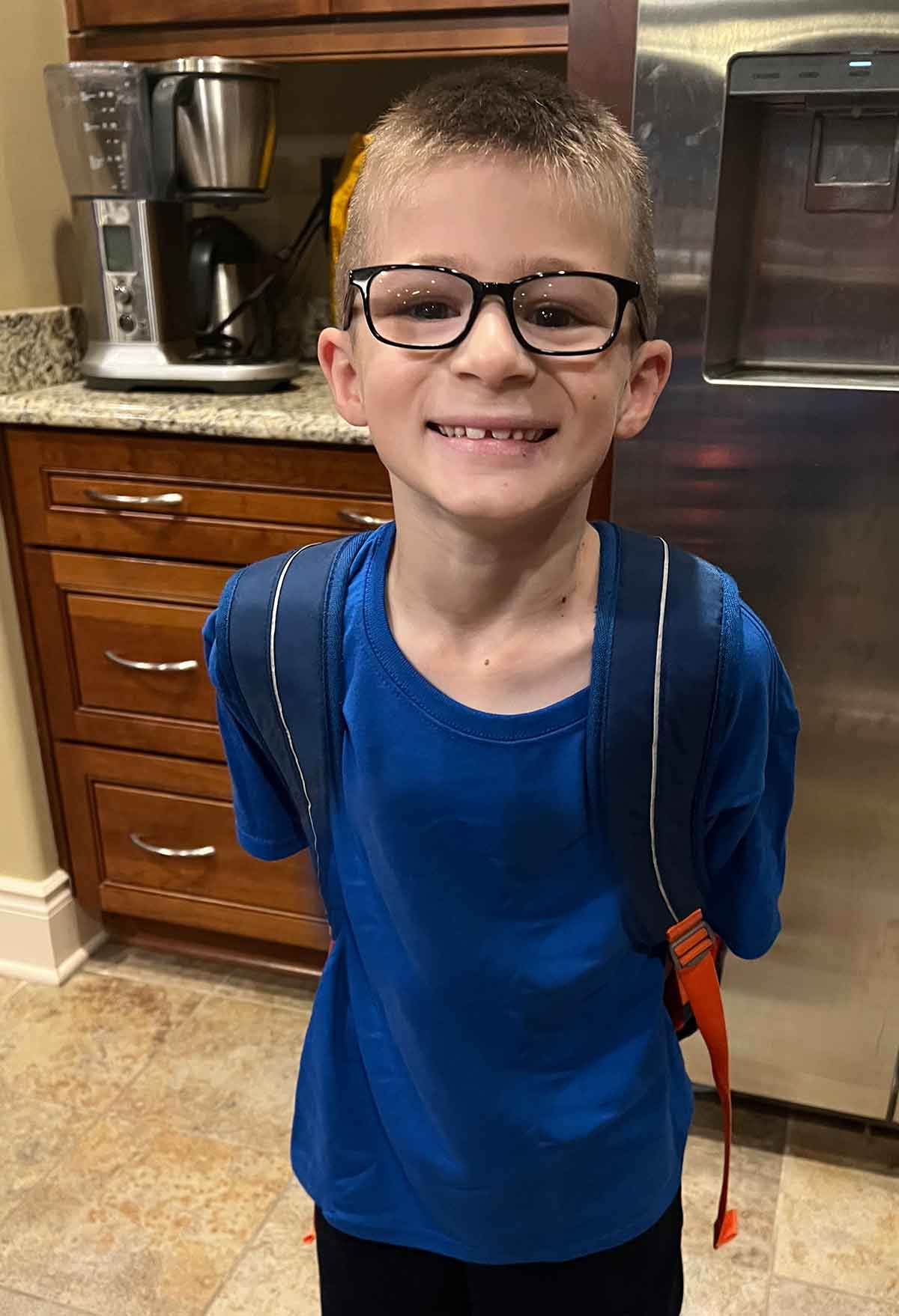 Boy in a blue tshirt and glasses with a backpack on standing in a kitchen.