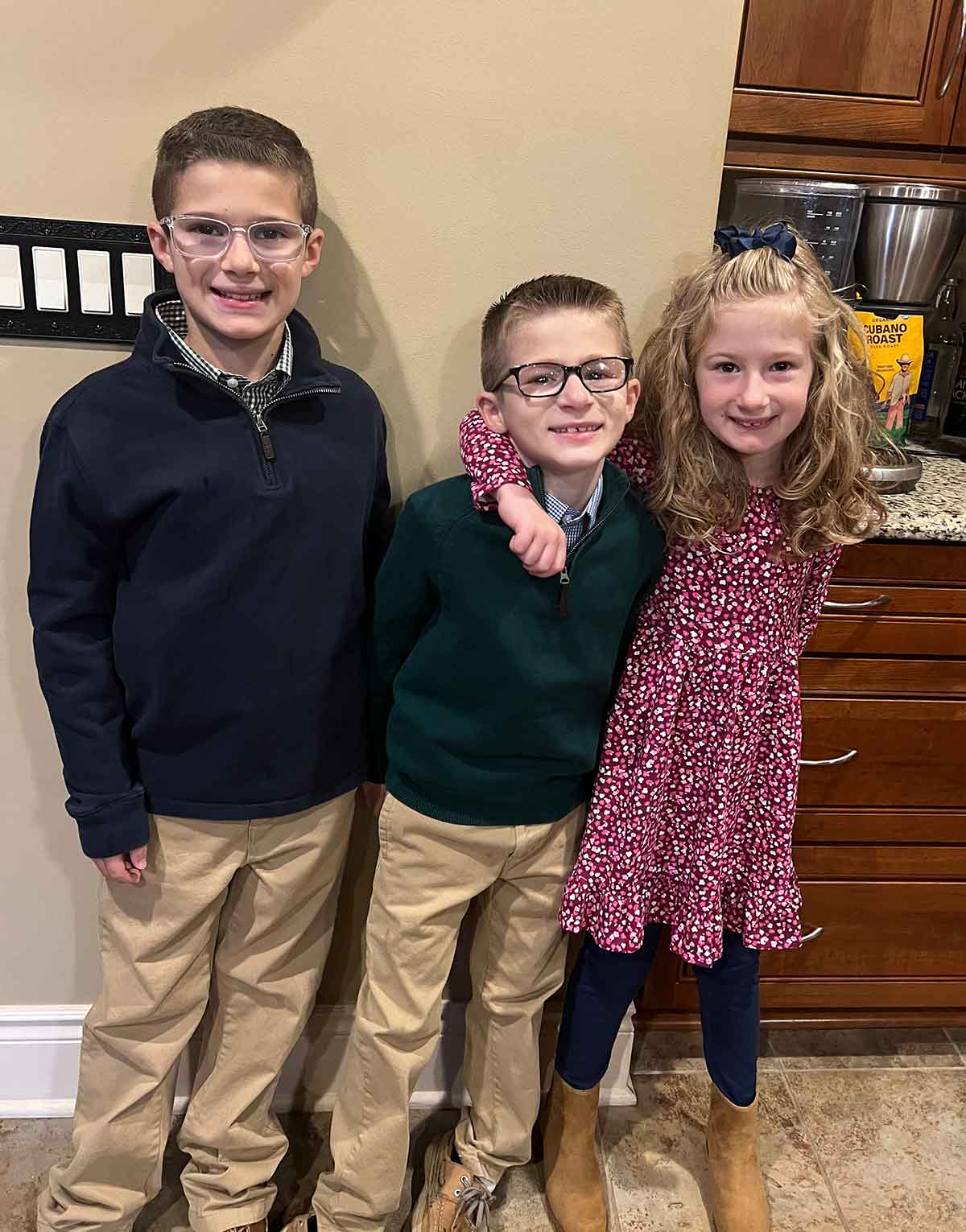 Two boys and a girl in dress clothes standing together in a kitchen.