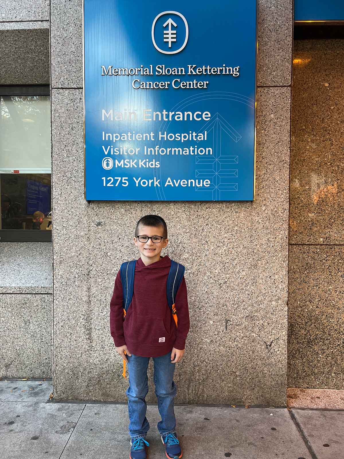 Boy with a backpack standing in front of a sign at the entrance to Memorial Sloan Kettering Cancer Center.