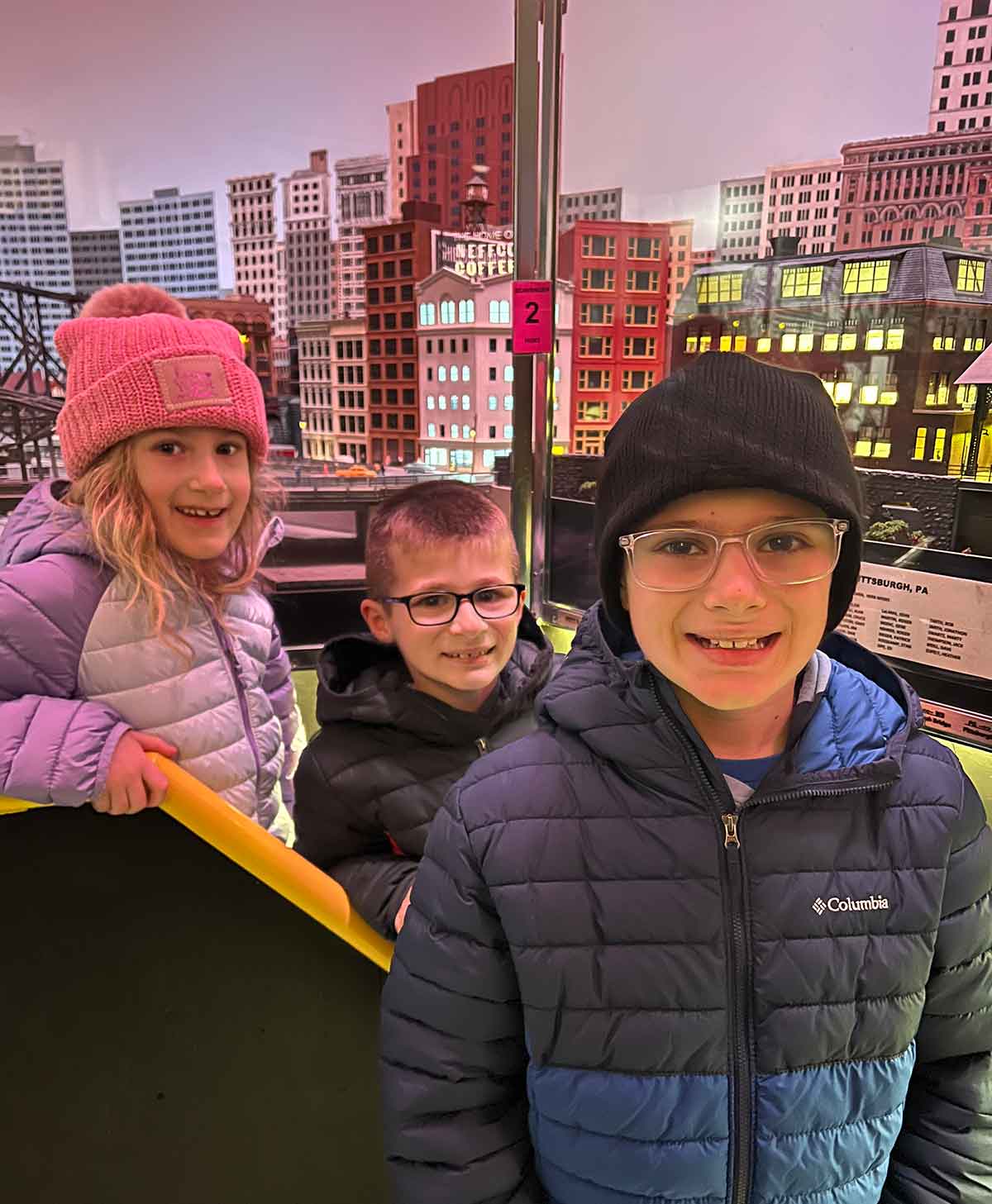 Three kids in winter jackets and hats standing in front of a model train display.