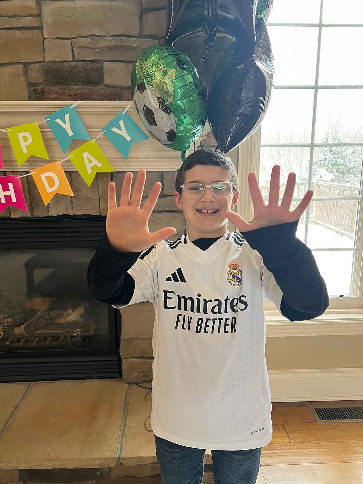 Boy in a soccer jersey standing in front of a birthday banner and balloons holding up ten fingers.