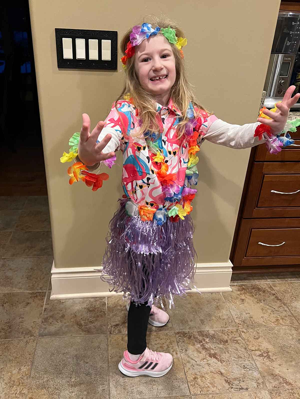 Blonde-haired little girl in a purple grass skirt, pink flamingo Hawaiian shirt, and leis around her neck, wrists, and in her hair.