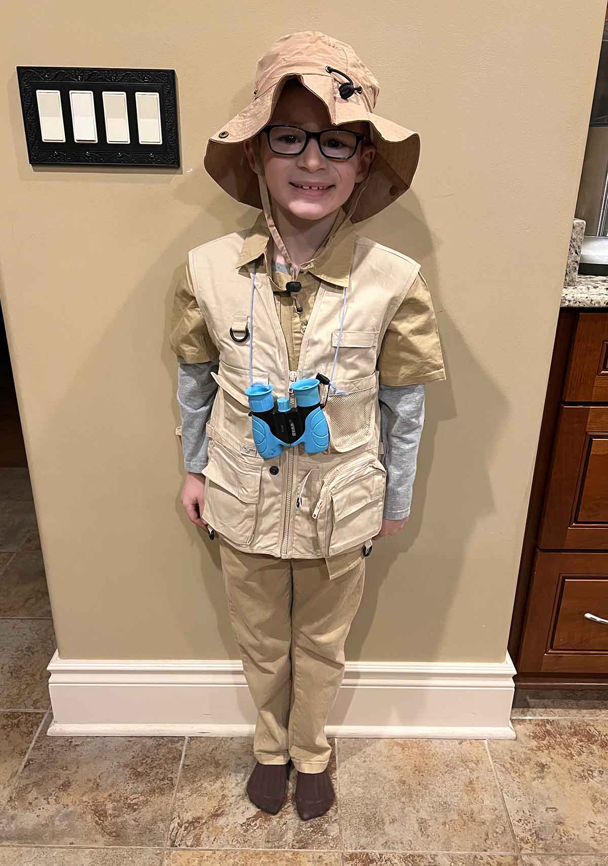 Little boy dressed in safari gear with khaki pants, khaki shirt, vest, and hat with binoculars around his neck.