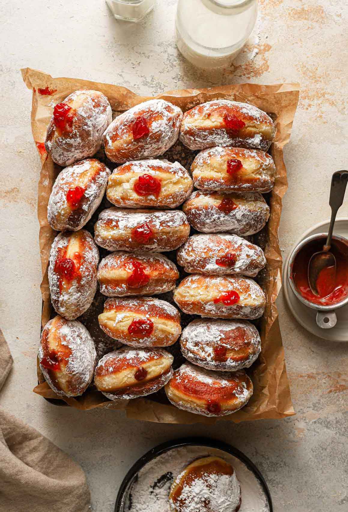 Overhead photo of paczki lined up on a parchment-lined baking sheet with a jar of milk, bowl of preserves, and powdered sugar surrounding it.