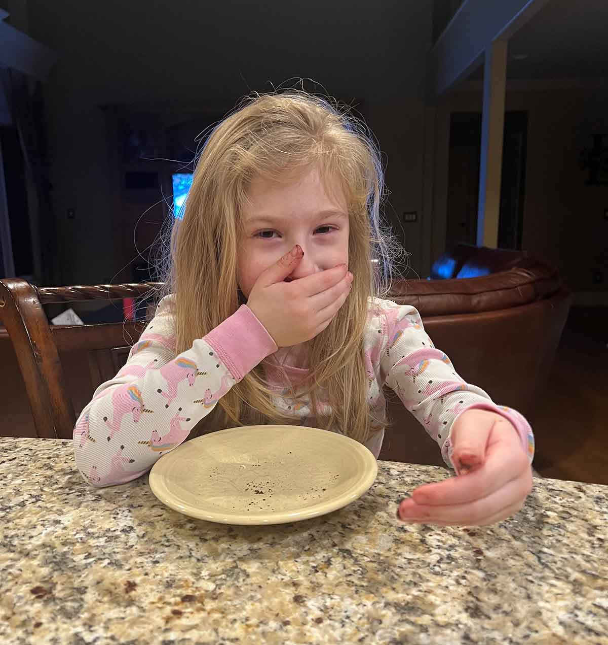 Little girl in pajamas sitting in front of an empty plate with a hand over her mouth.