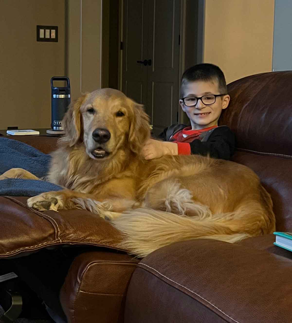 Boy in glasses sitting on a couch with a Golden Retriever dog.