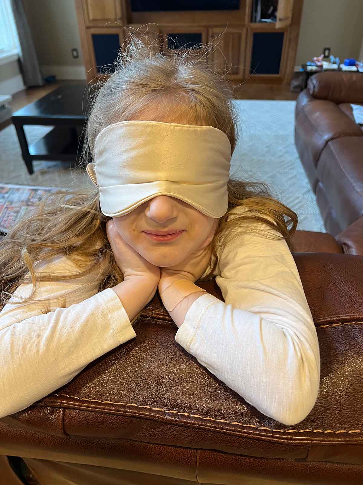 Little girl with an ivory sleep mask on, leaning over the back of a couch.